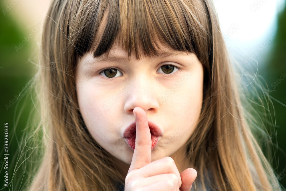 Naklejka premium Portrait of pretty child girl with gray eyes and long fair hair holding point finger on her lips with hush sigh. Cute female kid on warm summer day outside.