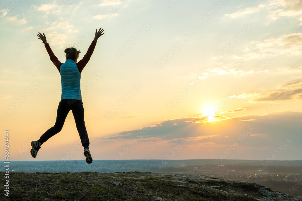 Silhouette of a woman hiker jumping alone on empty field at sunset in mountains. Female tourist raising her hands up in evening nature. Tourism, traveling and healthy lifestyle concept. Silhouette of a woman hiker jumping alone on empty field at sunset in mountains. Female tourist raising her hands up in evening nature. Tourism, traveling and healthy lifestyle concept.