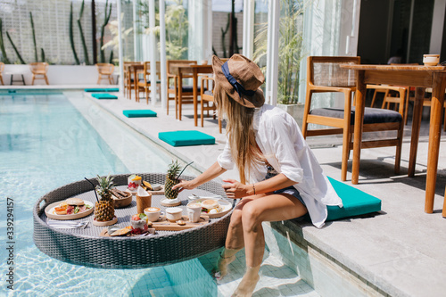 Slim girl in elegant brown hat eating juicy fruits at resort cafe. Graceful european woman in white shirt relaxing with cocktail and food in pool.