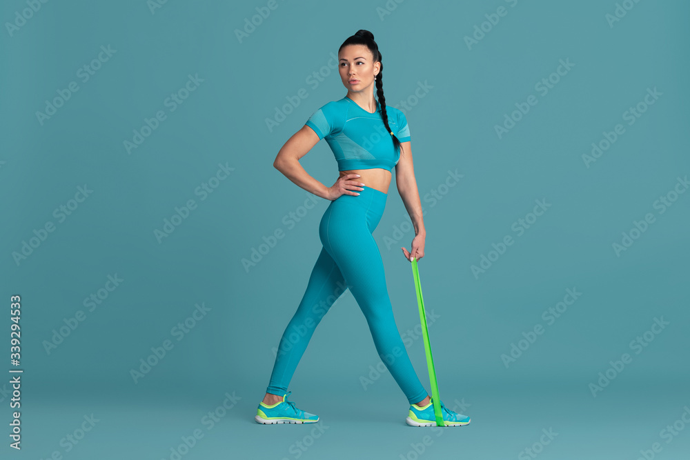 Fototapeta premium Legs and butt. Beautiful young female athlete practicing in studio, monochrome blue portrait. Sportive fit brunette model with elastics. Body building, healthy lifestyle, beauty and action concept.