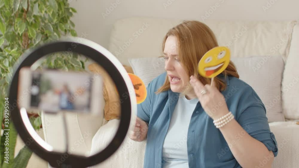 A Young Female Vlogger Recording a Video for an Online Blog with Smartphone and Ring Light. The Blogger Talks and Gestures and Uses Cardboard Signs with Hand Drawn Emoticons. Cozy White Home Interior. A Young Female Vlogger Recording a Video for an Online Blog with Smartphone and Ring Light. The Blogger Talks and Gestures and Uses Cardboard Signs with Hand Drawn Emoticons. Cozy White Home Interior.