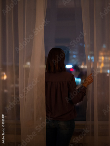Woman standing at window looking out at bright cityscape night lights