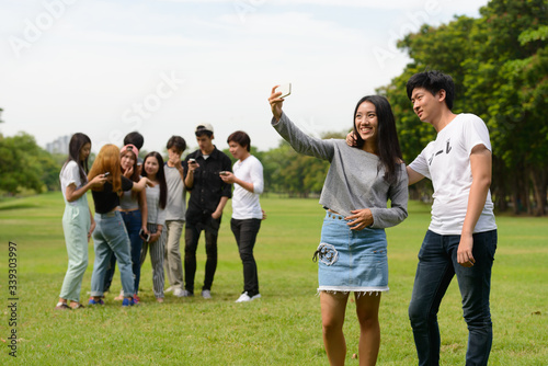 Happy young Asian couple taking selfie together with friends at the park