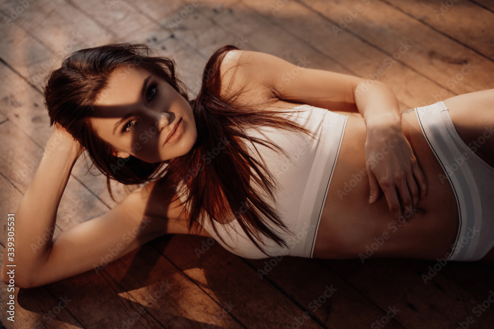 Obraz premium Almost naked and smooth fit woman posing in a bright apartment illuminated with sunlight, sitting on a white surface while wearing sporty underwear, looking lovely and erotic.