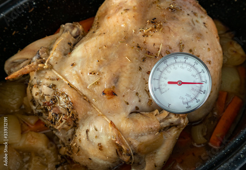 Measuring internal temperature of oven baked chicken. Meat / instant-read thermometer to measure food  safe temperature. Whole chicken in black pan with herb rub. Carrots and onions under the chicken. photo