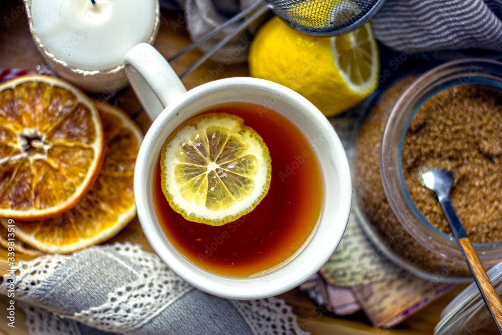 Hot black tea with a slice of lemon in a large gray mug with a spoon closeup, top view, next to dried oranges, fresh lemon, brown sugar, gray napkin on a wooden tray. The concept of a cozy home tea Hot black tea with a slice of lemon in a large gray mug with a spoon closeup, top view, next to dried oranges, fresh lemon, brown sugar, gray napkin on a wooden tray. The concept of a cozy home tea