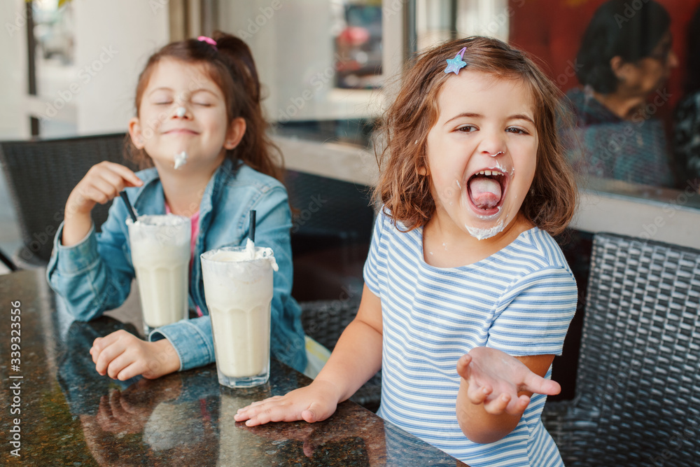 Two funny Caucasian little preschool sisters siblings drink milk shakes in  a cafe. Friends girls having fun together. Cold summer desserts for kids.  Happy authentic childhood lifestyle. Photos | Adobe Stock