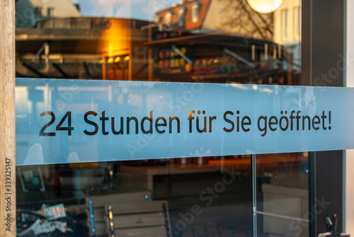 Schaufenster von Ladengeschäft mit Schild „24 Stunden für Sie geöffnet“, in Deutsch, mit Auslagen und die Spiegelung der Umgebung in Unschärfe zur Zeit der Corona Covid-19 Pandemie im März 2020