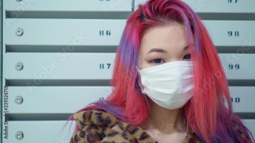 Concept: urban healthcare during pollution pandemic. Beautiful young asian girl colored hair medical mask grunge fashion clothing standing near mailboxes in apartment house