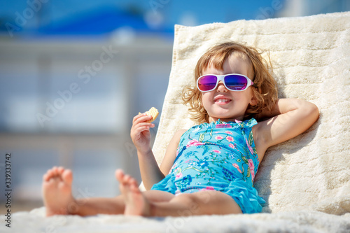 Adorable little girl enjoying on a sun lounger rest on the beach chair on tropical sandy beach sea shore. Sunbathing and leisure on sunny day. Travel with young children