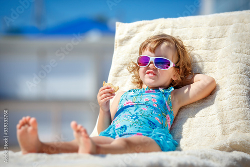 Adorable little girl enjoying on a sun lounger rest on the beach chair on tropical sandy beach sea shore. Sunbathing and leisure on sunny day. Travel with young children