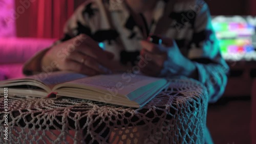 man flips through book.He is holding a pipe and a glass of tea.cozy and warm, he sits in front of the table and reads is dressed in pyjamas has stubble mustache.the room has colored lights pink blue