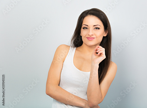 Beautiful positive young happy woman with hand under the face thinking and looking in casual white t-shirt and long hair on blue background. Closeup