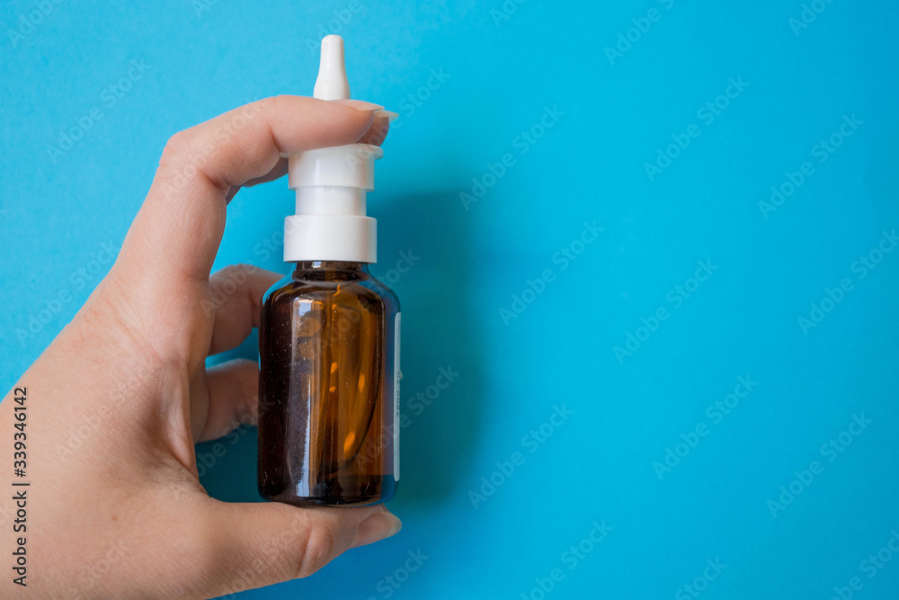 Fototapeta premium Nasal spray with sea water for the treatment and prevention of SARS symptoms. A glass bottle of sea water with a nozzle for irrigation of the nasal mucosa in a woman's hand on a blue background with K