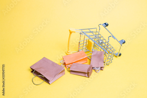 overturned miniature shopping trolley on a yellow background, fallen small paper bags, copy space, online shopping concept