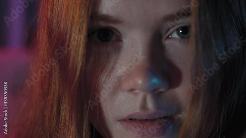 portrait of a cute red-haired girl looking at the camera.the woman has blue eyes and red hair.the room has dark lighting pink and blue