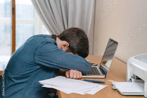 Burnout young man is crying in his home office, sitting at his desk with a laptop and documents. Tired depressed freelancer frustrated by business failure, feel exhausted, worried about work problems