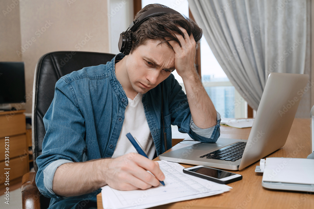 Exausted young bussinessman working hard on new progect at home office, using laptop, writting on papers. Bored university student wearing headphones tired of distance learning. Remote work concept Exausted young bussinessman working hard on new progect at home office, using laptop, writting on papers. Bored university student wearing headphones tired of distance learning. Remote work concept
