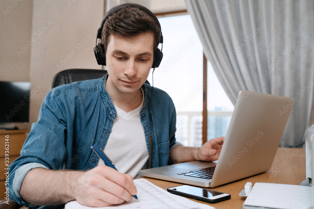 Happy young man in headphones sitting at desk in home office, writting on papers, using laptop for distance education, doing homework, writing notes, listerning audio course, watching webinar Happy young man in headphones sitting at desk in home office, writting on papers, using laptop for distance education, doing homework, writing notes, listerning audio course, watching webinar