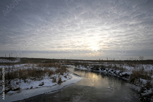A sunset sky of quilted clouds over a quiet stream flowing through a prairie landscape in winter.