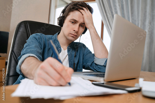 Tired young man working at home office on new project, frustrated guy sitting at his desk writing on papers, thinking on difficult task. Bored student studying from home, doing home work. E-learning