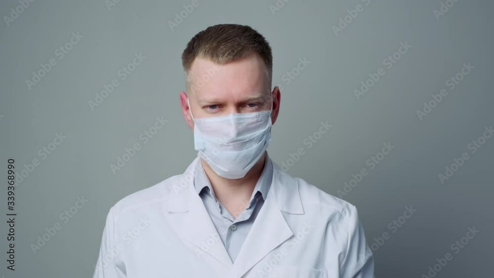A young man doctor in a mask and a white robe on a gray background. Putting on a mask. A young man doctor in a mask and a white robe on a gray background. Putting on a mask.