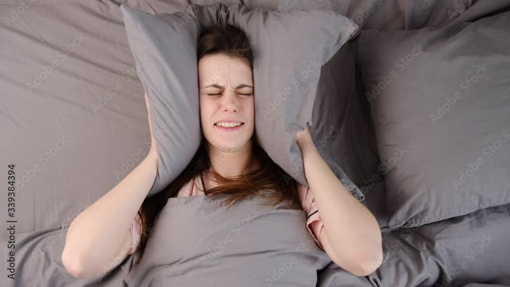 Woman lying in bed covering head with pillow because too loud annoying noise keeps her up all night long. Irritated girl disturbed with loud noise problem wake up lying in bed suffer from insomnia Woman lying in bed covering head with pillow because too loud annoying noise keeps her up all night long. Irritated girl disturbed with loud noise problem wake up lying in bed suffer from insomnia