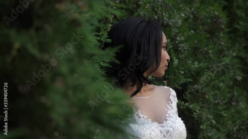 elegant African american bride wearing beautiful white dress with laces poses half face among bushes closeup slow motion photo