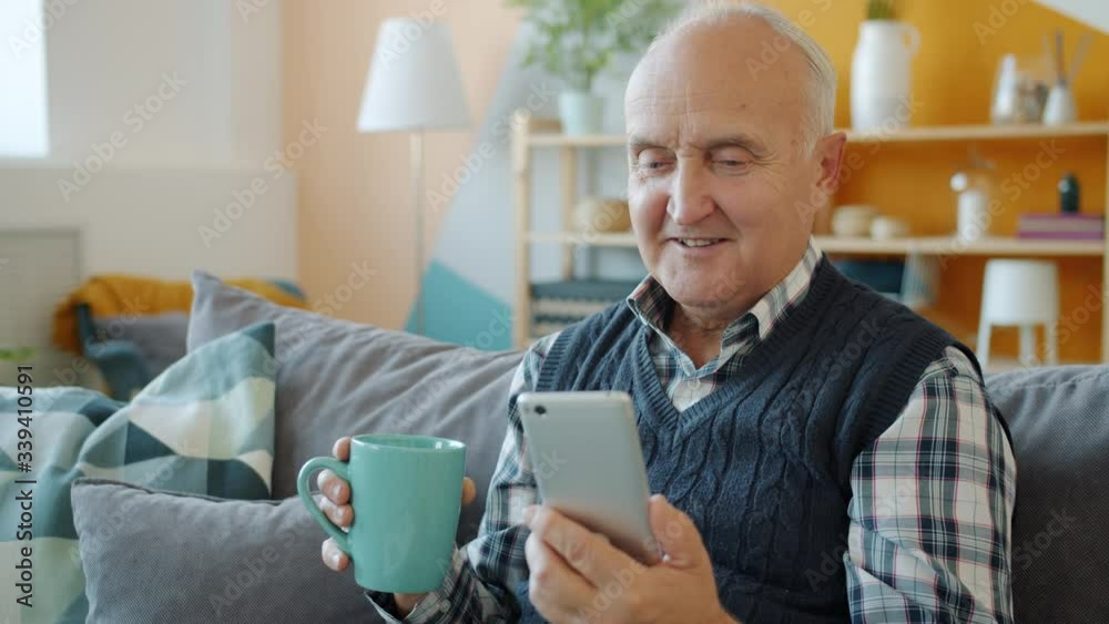 Elderly man is watching content on smartphone screen and drinking tea relaxing at home smiling sitting on couch alone. Devices and retired people concept. Elderly man is watching content on smartphone screen and drinking tea relaxing at home smiling sitting on couch alone. Devices and retired people concept.