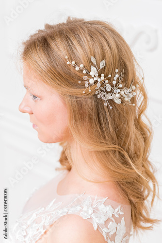 Romantic shy young bride in elegant hotel room in white lace wedding dress with makeup and wavy blonde hair with pearl jewelry getting ready in bride's morning. Fine art wedding. 