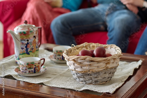 Easter eggs in basket on table with tea pot and tea cups - unrecognizable people sitting on sofa