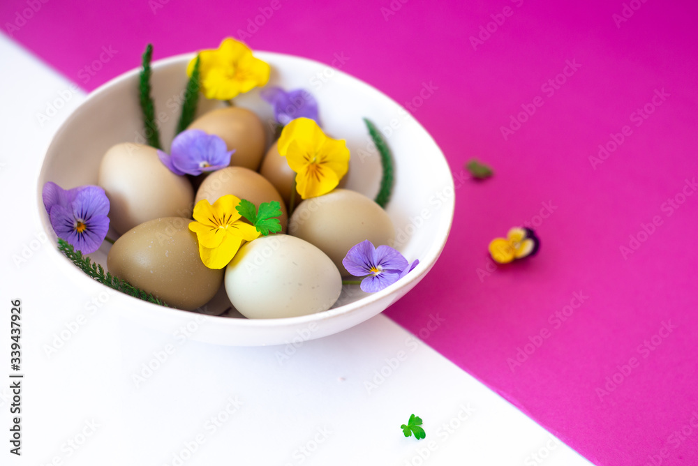 Easter eggs with yellow and lilac flowers in a white bowl on a white and pink background. Easter concept, copy text. Easter eggs with yellow and lilac flowers in a white bowl on a white and pink background. Easter concept, copy text.