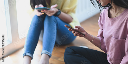 Cropped image of beautiful women relaxing by using their smartphone while sitting together at the floor over comfortable sitting room as background.