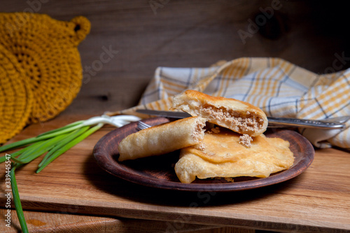 Clay plate of fried meat pies with cutlery and green onion on wooden table.