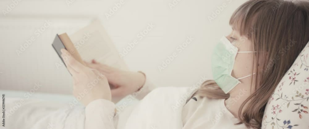 Woman with surgical face mask reading a book in bed staying home during coronavirus covid 19 virus outbreak close up from behind Woman with surgical face mask reading a book in bed staying home during coronavirus covid 19 virus outbreak close up from behind