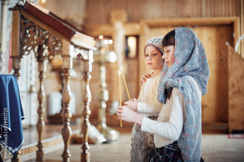 two Russian girls in a scarf on their heads stand in an Orthodox Church, lights a candle and prays in front of the icon.