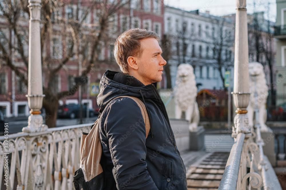 A young blond man in a jacket and with a backpack stands on the bridge with statues of white lions in the middle of the canals of St. Petersburg, Attractions, morning spring