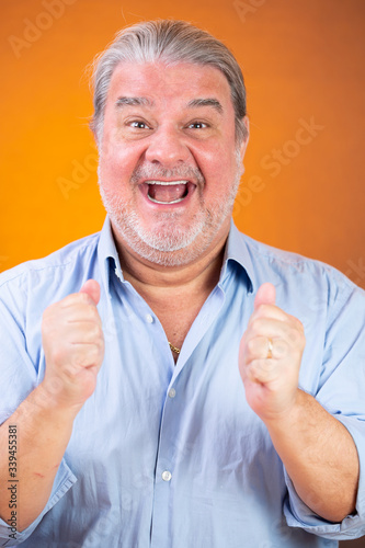 Man headshot portrait. He is happy. He is smiling to the camera and has her two hands showing thumps up like saying good work. The background is lighted with orange color.