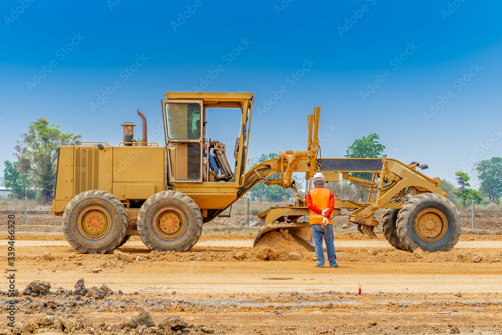 Motor grader clearing and leveling construction site surface while worker controlling level point. Grader industrial machine on road construction work.
