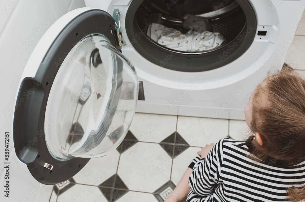 Cute girl in striped clothes puts things into the washing machine. Cleanliness and Household Concept Cute girl in striped clothes puts things into the washing machine. Cleanliness and Household Concept