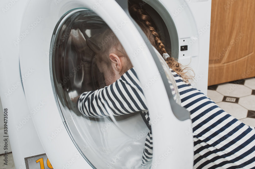 Cute girl in striped clothes puts things into the washing machine. Cleanliness and Household Concept