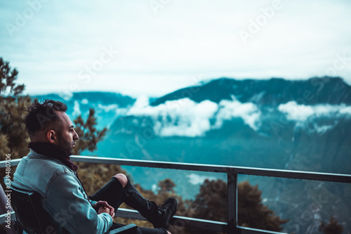 Armenian Caucasian traveler sitting on a terrace surrounded by mountains in the early morning. Cloudy and foggy sky at the dawn. Breathing fresh air and drinking hot tea from mug. Travel concept.