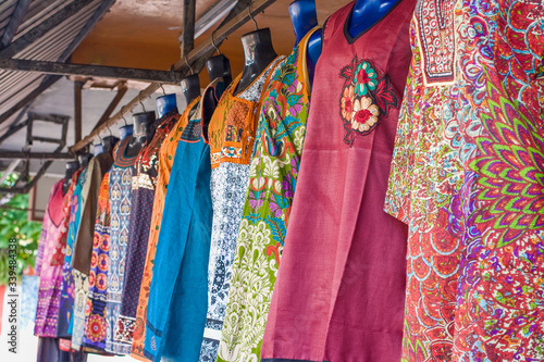Brightly coloured cotton womens dresses for sale, hanging from a metal rack at covered street market stall Goodlands, Mauritius during summer.