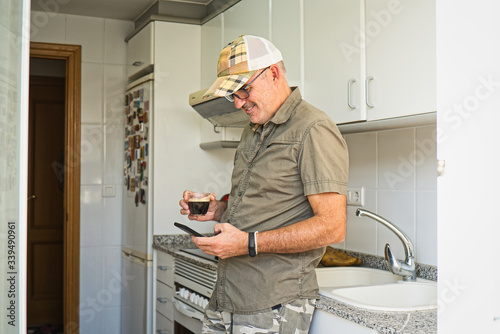 Adult Western male with glasses, cap and activity bracelet drinking espresso standing in a modern white home kitchen. Green summer clothes. Checking his smartphone, smiling. 50´s. Magnets.