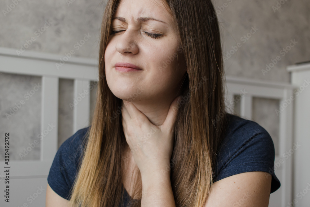 Young unhappy woman has sore throat. The girl feels pain, symptoms of the beginning of a cold, flu or virus. Tired girl sitting at home having pain in the neck Young unhappy woman has sore throat. The girl feels pain, symptoms of the beginning of a cold, flu or virus. Tired girl sitting at home having pain in the neck