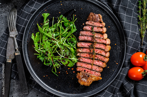 Cooked grilled strip loin steak, marbled beef meat with arugula. Black background. Top view