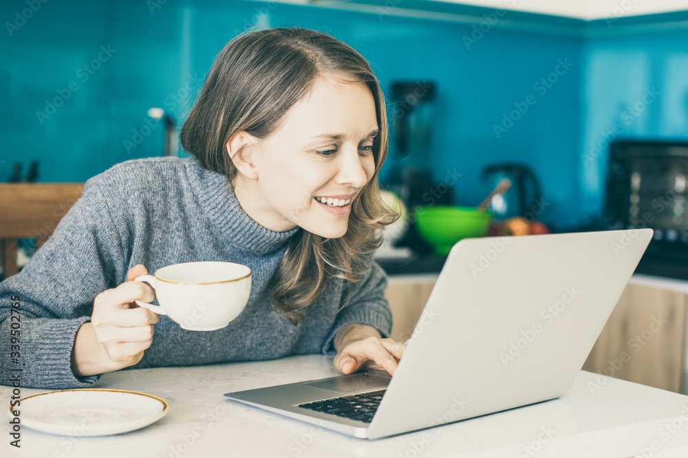 Closeup portrait of focused young beautiful woman drinking coffee and browsing on laptop computer in kitchen. Freelancer concept.