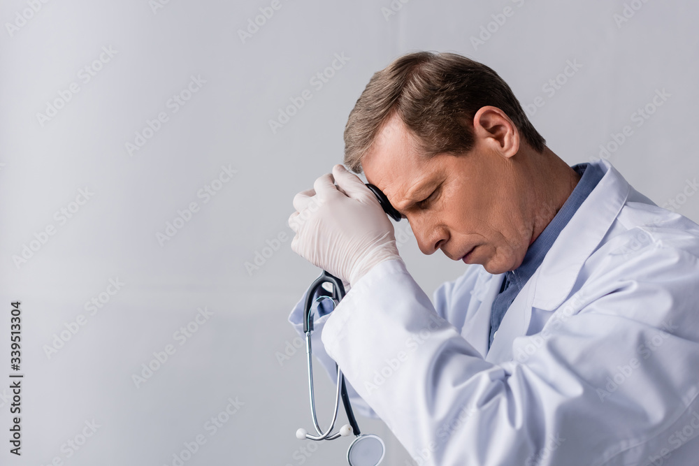 tired and mature doctor with closed eyes holding stethoscope on grey