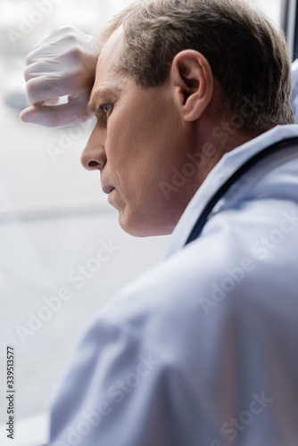 selective focus of tired middle aged doctor standing near window in clinic