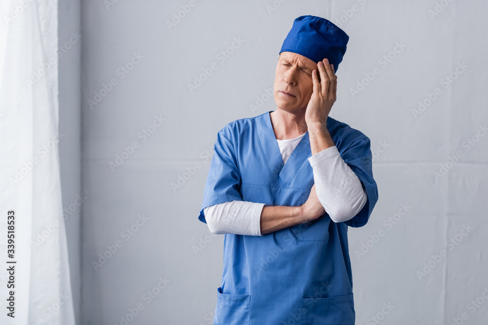 tired and middle aged doctor in blue scrub hat touching head on grey